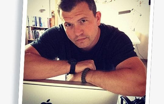 Man sitting with folded arms on an Apple computer, room with books and desk lamp, wearing watch.