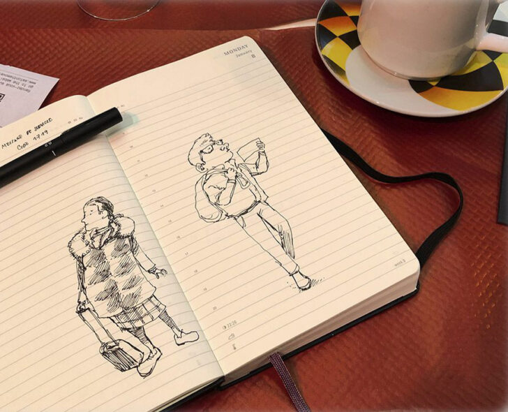 Pen doodles of unsuspecting train station travelers in sketchbook beside a cup of coffee and croissant on table.