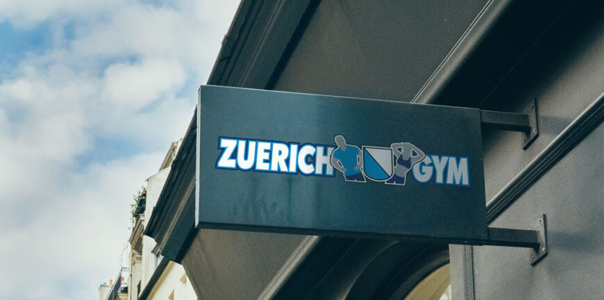 Outdoor sign for Zuerich Gym with stylized athletes and a shield on a building exterior.