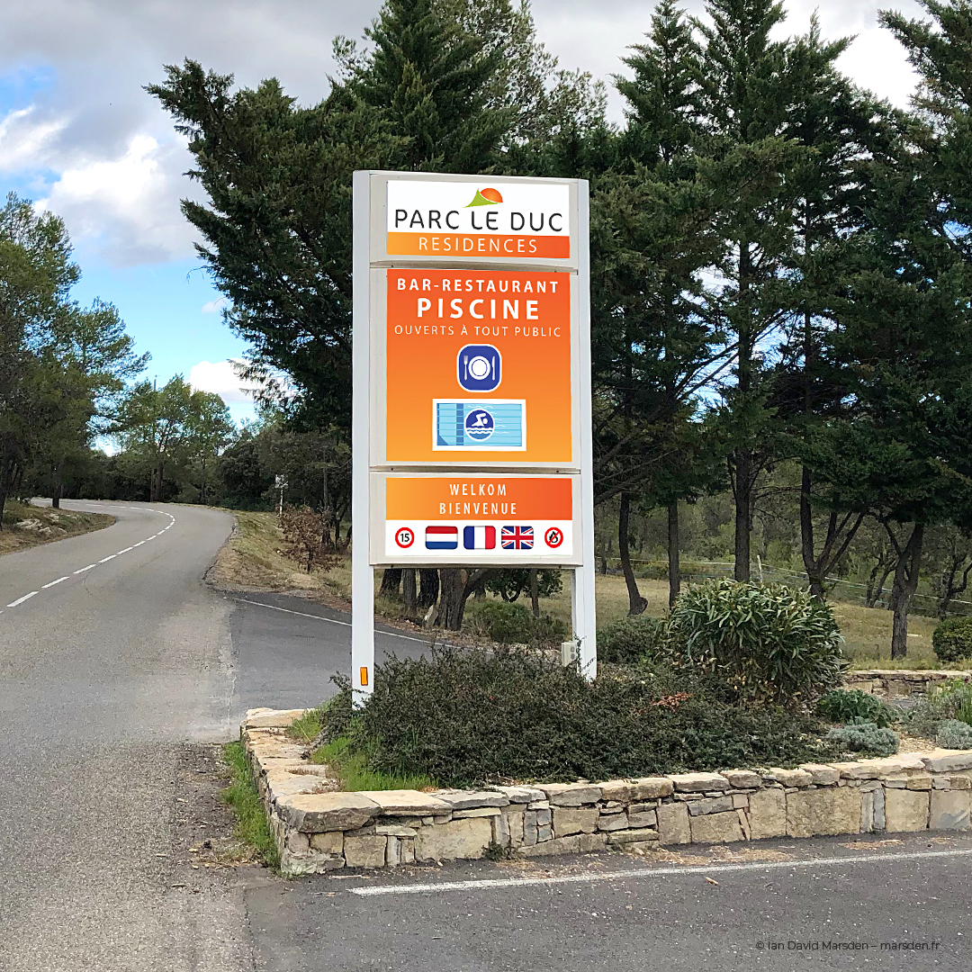 Main entrance sign for Parc Le Duc with multilingual welcome and pool/restaurant visuals, designed for roadside visibility on a curve.