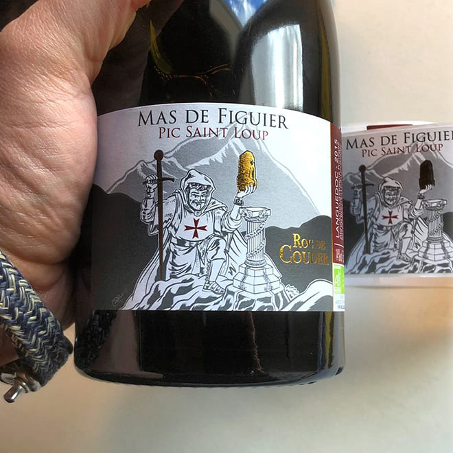 Bouteille Roc de Couder avec étiquette finale posée, devant un rouleau d’étiquettes imprimées pour le Mas de Figuier.