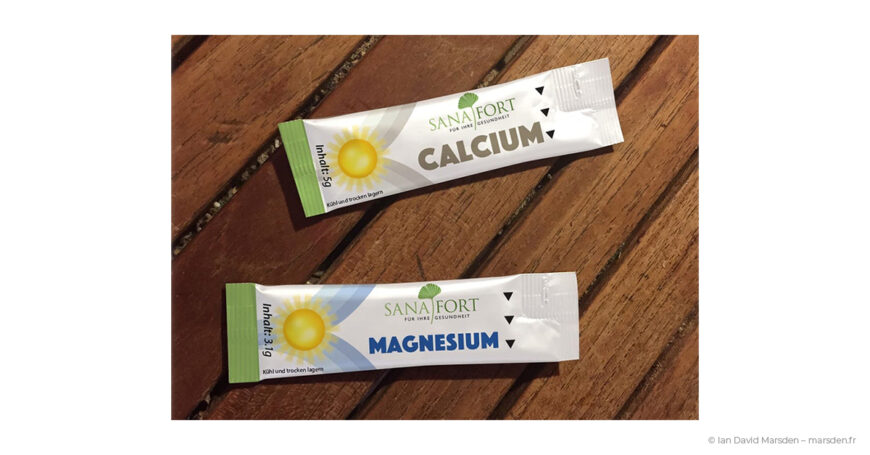 Photograph of Sanafort Calcium and Magnesium single-serve stick sachets designed by Ian David Marsden, shown on a wooden surface with coordinated product colors and the Sanafort logo.