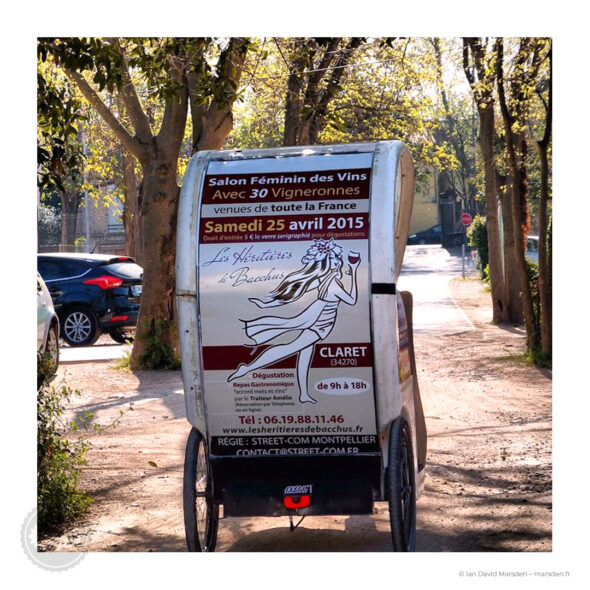 Vue arrière d’un vélo-taxi publicitaire montrant la grande illustration féminine des Héritières de Bacchus et les informations du salon 2015 à Claret.