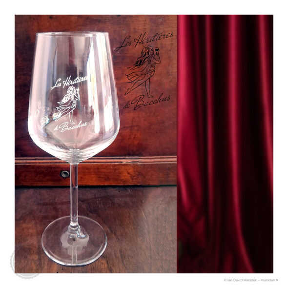 Verre à vin transparent sérigraphié du logo Les Héritières de Bacchus, photographié sur fond de bois avec un rideau rouge.