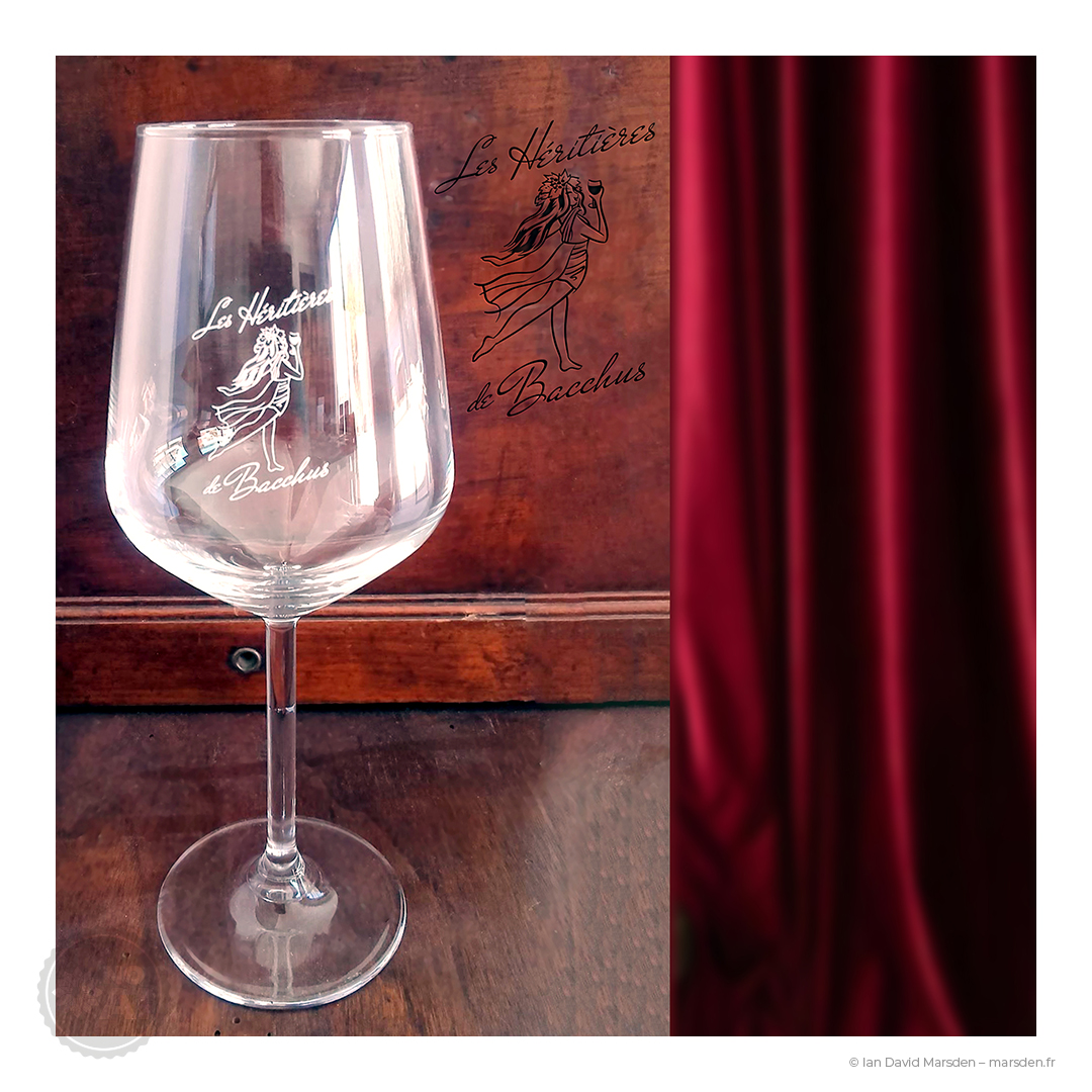 Verre à vin transparent sérigraphié du logo Les Héritières de Bacchus, photographié sur fond de bois avec un rideau rouge.
